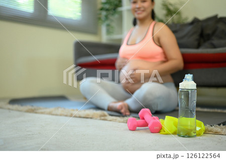 Pregnant woman sitting on the floor with a bottle of water and two dumbbells in front of her 126122564
