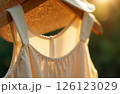 Women's beige sundress and straw hat hanging on wooden hanger on green background, glare of sun 126123029