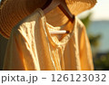 Woman Shirt Dress And Straw Hat Hanging On Hanger In Sun In Background In Blur Sea And Greenery 126123032