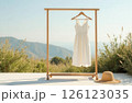 A white dress hangs on a wooden clothes hanger, a straw hat lies on the floor, against the background of nature 126123035