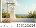 A white woman's dress hangs on a hanger, a straw hat lies on the floor next to it on a white terrace 126123036