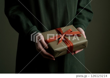 Close-up of a person in a dark green sweater holding an elegant gift wrapped in brown paper with a red ribbon. Minimalist style, soft lighting, warm and festive atmosphere. Copy space. Banner Close-up of a person in a dark green sweater holding an elegant gift wrapped in brown paper with a red ribbon. Minimalist style, soft lighting, warm and festive atmosphere. Copy space. Banner 126123214