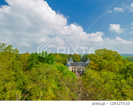 Aerial view of a historic castle surrounded by a forest, with vast green fields, winding waterways, and a river stretching across the flat Dutch countryside under a partly cloudy sky. 126125510