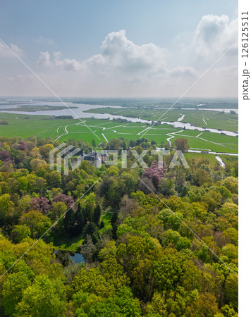 Aerial view of a historic castle surrounded by a forest, with vast green fields, winding waterways, and a river stretching across the flat Dutch countryside under a partly cloudy sky. 126125511