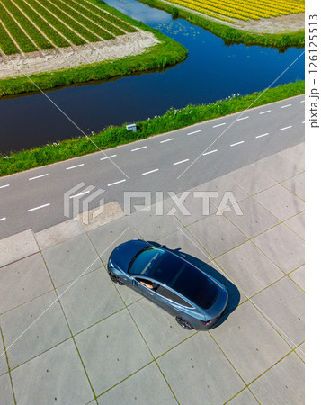 Aerial view of a single parked car on a large empty concrete lot next to a road and canal, with green grass and clear road markings under bright daylight. 126125513