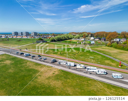 Aerial view of a green open field with a row of camper vans parked along a road, modern and traditional houses in the background, under a bright blue sky with scattered clouds. 126125514