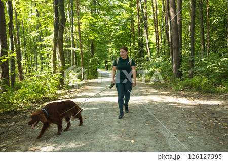 Woman Walking with Irish Setter Dog on Forest Trail Surrounded by Lush Green Trees 126127395