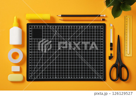 A black cutting mat with white grid lines sits on a bright yellow background surrounded by neatly laid out office supplies: pencils, glue, tape, scissors, cutter, erasers and ruler. 126129527