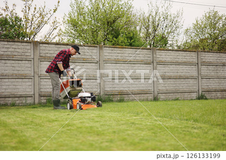 man in casual clothes holds bag with cut grass of lawn mower after mowing lawn with at own backyard. Husband takes care of garden. Modern gasoline garden equipment. Landscaping work at home 126133199