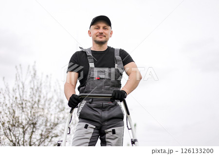 Bottom view of professional male gardener in protective apparel, gloves, cap and overalls holds handle of gasoline cordless lawnmower for mowing grass lawn outdoors. Seasonal landscaping design work 126133200
