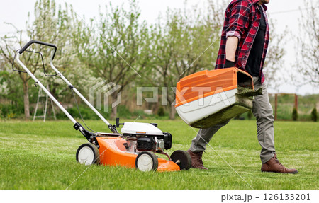 man in casual clothes holds bag with cut grass of lawn mower after mowing lawn with at own backyard. Husband takes care of garden. Modern gasoline garden equipment. Landscaping work at home 126133201