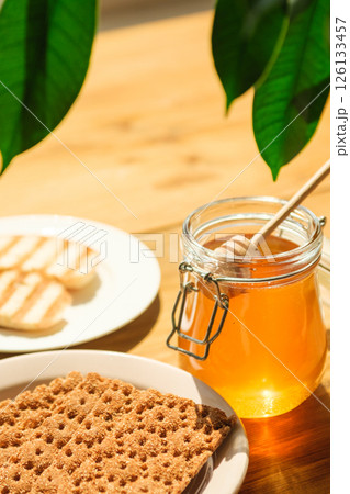 Honey Jar and Wooden Dipper Next to Crispbread and Cookies on a Sunny Wooden Table, Evoking Cafes and Wellness Food Honey Jar and Wooden Dipper Next to Crispbread and Cookies on a Sunny Wooden Table, Evoking Cafes and Wellness Food 126133457