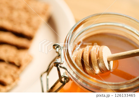 A Glass Jar Filled With Golden Honey Sits With a Wooden Dipper, Accompanied by Crisp Crackers on a Beige Plate in a Cozy Kitchen A Glass Jar Filled With Golden Honey Sits With a Wooden Dipper, Accompanied by Crisp Crackers on a Beige Plate in a Cozy Kitchen 126133465