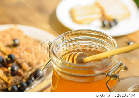 A Glass Jar Filled With Golden Honey Sits on a Rustic Table, With a Wooden Stick for Stirring Alongside Cookies and Fresh Berries A Glass Jar Filled With Golden Honey Sits on a Rustic Table, With a Wooden Stick for Stirring Alongside Cookies and Fresh Berries 126133474