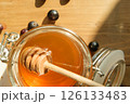 Glorious Honey Jar Filled With Golden Honey and a Wooden Dipper Sits on a Sunlit Wooden Table, Surrounded by Dark Berries 126133483