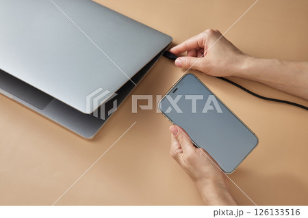 female hand connecting charger cable to laptop and holding smartphone at beige table, closeup female hand connecting charger cable to laptop and holding smartphone at beige table, closeup 126133516