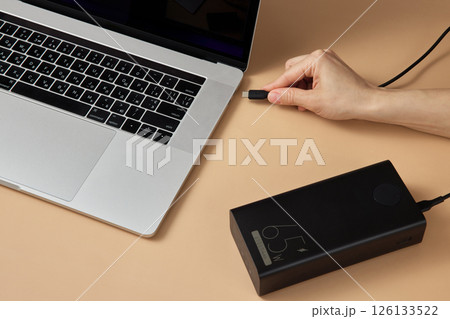female hand connecting charger cable of power bank to laptop on beige background, close-up. 126133522
