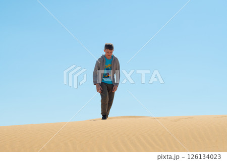 Boy walking on top of a sand dune in the Kyzylkum Desert under a bright blue sky. The golden ripples of sand evoke a sense of freedom and exploration Boy walking on top of a sand dune in the Kyzylkum Desert under a bright blue sky. The golden ripples of sand evoke a sense of freedom and exploration 126134023