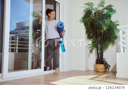Woman Holding Blue Yoga Mat and Water Bottle Standing in Bright Room With Indoor Plants Woman Holding Blue Yoga Mat and Water Bottle Standing in Bright Room With Indoor Plants 126134074