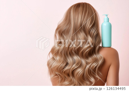 A woman displaying her long, wavy, blonde hair with a focus on a blue shampoo bottle, illustrating clean and healthy hair care enhancing beauty. 126134495