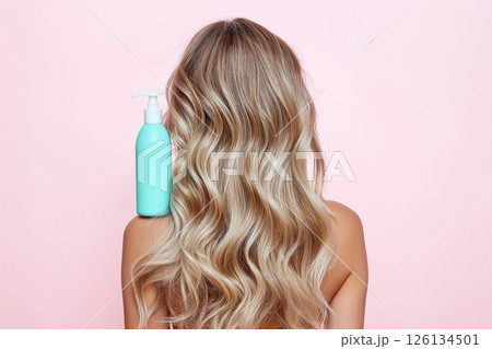 A woman displaying her long, wavy, blonde hair with a focus on a blue shampoo bottle, illustrating clean and healthy hair care enhancing beauty. 126134501