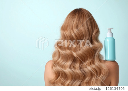 A woman displaying her long, wavy, blonde hair with a focus on a blue shampoo bottle, illustrating clean and healthy hair care enhancing beauty. 126134503