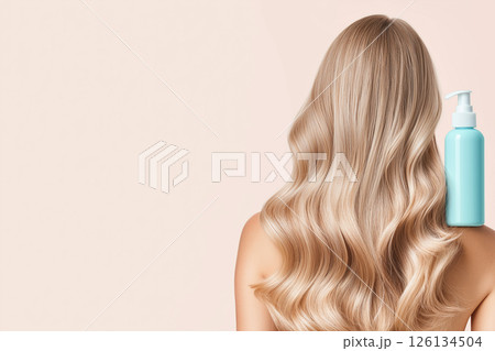 A woman displaying her long, wavy, blonde hair with a focus on a blue shampoo bottle, illustrating clean and healthy hair care enhancing beauty. 126134504