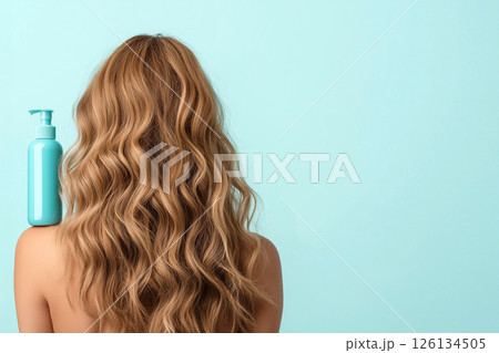A woman displaying her long, wavy, blonde hair with a focus on a blue shampoo bottle, illustrating clean and healthy hair care enhancing beauty. 126134505