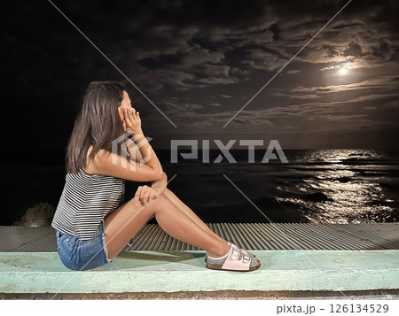 young woman sits at night on the seashore and dreamily looks at the moon in the sky young woman sits at night on the seashore and dreamily looks at the moon in the sky 126134529