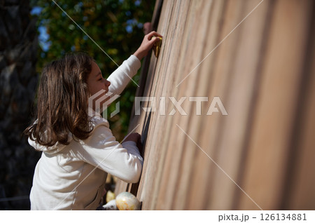 Young Girl Enthusiastically Climbing a Wooden Wall Outdoors Young Girl Enthusiastically Climbing a Wooden Wall Outdoors 126134881