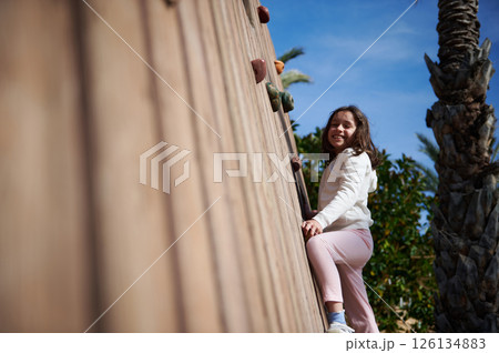 Joyful Girl Climbing Outdoor Wooden Wall on a Bright Day Joyful Girl Climbing Outdoor Wooden Wall on a Bright Day 126134883