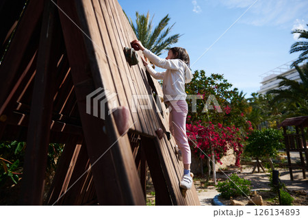 Young Girl Climbing a Wooden Wall in a Children's Outdoor Playground 126134893