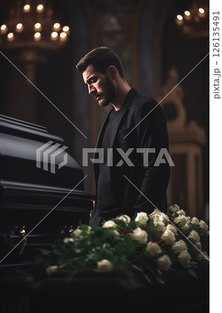 Young Caucasian man says goodbye to a dead man in a church. A coffin, flowers and a grieving relative are nearby. Vertical photo. 126135491