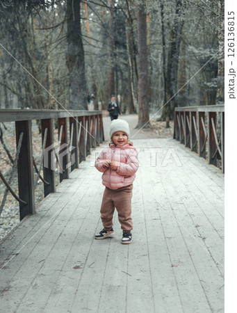 Happy child in pink jacket and hat standing on wooden bridge surrounded by trees in serene winter forest enjoying leisurely walk among beauty of nature. Cute little kid on walk in park. Happy child in pink jacket and hat standing on wooden bridge surrounded by trees in serene winter forest enjoying leisurely walk among beauty of nature. Cute little kid on walk in park. 126136815