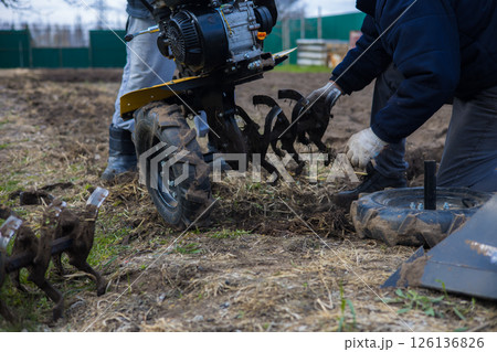 A farmer installs a cultivator on a walk-behind tractor, prepares to plow the land and cultivate the field, demonstrating land preparation techniques. Plowing the land with a walk-behind tractor. A farmer installs a cultivator on a walk-behind tractor, prepares to plow the land and cultivate the field, demonstrating land preparation techniques. Plowing the land with a walk-behind tractor. 126136826