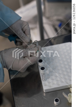 A veterinarian places a piece of tissue in a test tube. A veterinarian wearing gloves and a lab coat prepares samples for analysis in a veterinary lab, holding the test tube and using instruments. A veterinarian places a piece of tissue in a test tube. A veterinarian wearing gloves and a lab coat prepares samples for analysis in a veterinary lab, holding the test tube and using instruments. 126136856