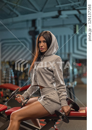 Young woman in a gray workout outfit sits confidently on a bench in a modern gym holding dumbbells while focused on her fitness routine. The gym features bright lighting and equipment. 126137168