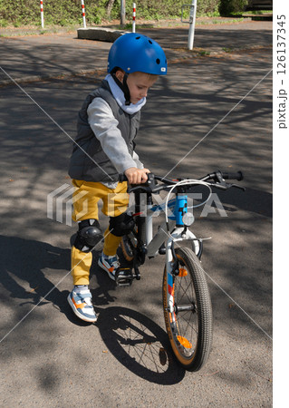 Child wearing safety gear learning to ride a bicycle in a park. Focused, determined expression while pushing bike along path. Bright helmet and knee pads for protection. 126137345