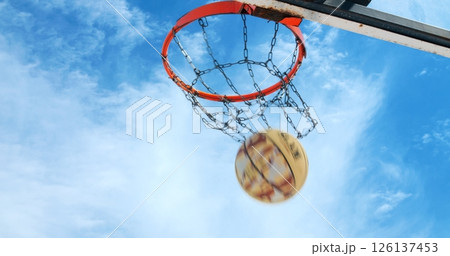 Basketball swishing through metal chain net, arcing against blue clouded sky during outdoor recreational game moment 126137453