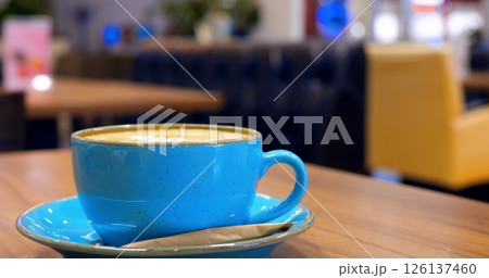 Professional barista carefully stirring creamy cappuccino in elegant blue ceramic cup, resting on white saucer atop rustic wooden cafe surface, showcasing intricate coffee preparation technique 126137460