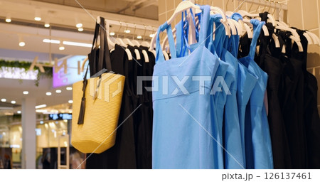 Stylish blue sundresses and black dresses hanging on clothing rack with yellow tote bag, showcasing contemporary fashion collection inside brightly illuminated retail space. Concept fashion clothes 126137461