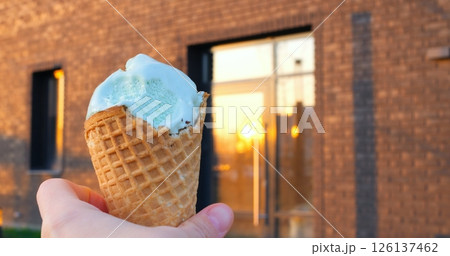 Concept of street sweets, ice cream in city. Mint ice cream scoop nestled in crispy waffle cone, held against weathered brick wall with reflective urban windows, catching warm sunset glow 126137462