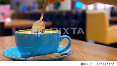 Professional barista carefully stirring creamy cappuccino in elegant blue ceramic cup, resting on white saucer atop rustic wooden cafe surface, showcasing intricate coffee preparation technique Professional barista carefully stirring creamy cappuccino in elegant blue ceramic cup, resting on white saucer atop rustic wooden cafe surface, showcasing intricate coffee preparation technique 126137472