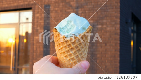 Mint ice cream scoop nestled in crispy waffle cone, held against weathered brick wall with reflective urban windows, catching warm sunset glow 126137507