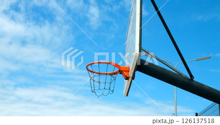 Green basketball swishing through orange hoop, arcing against blue sky with scattered clouds, showcasing athletic precision and scoring dynamics of streetball sport 126137518