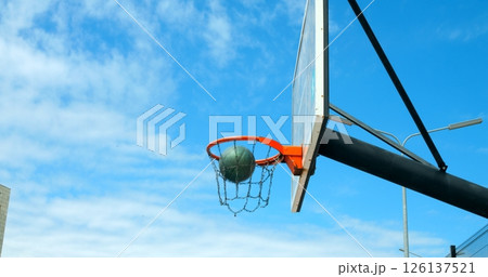 Basketball swishing through metal chain net, arcing against blue clouded sky during outdoor recreational game moment Basketball swishing through metal chain net, arcing against blue clouded sky during outdoor recreational game moment 126137521