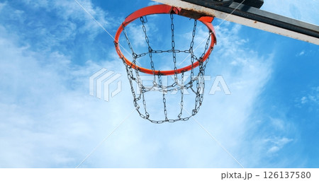 Basketball swishing through metal chain net, arcing against blue clouded sky during outdoor recreational game moment 126137580