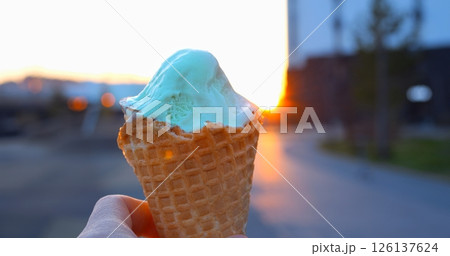 Creamy light green pistachio ice cream nestled in waffle cone, gleaming against blurred urban sunset street with golden evening glow and soft bokeh background Creamy light green pistachio ice cream nestled in waffle cone, gleaming against blurred urban sunset street with golden evening glow and soft bokeh background 126137624