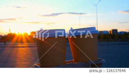 Sunlight shines between two disposable coffee cups on a cardboard tray, creating a starburst effect, with a blurred urban background at sunset 126137646