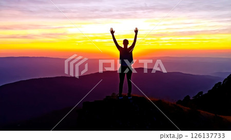 Hiker standing with raised arms on mountain peak, silhouetted against vibrant sunset, experiencing moment of triumph and serenity in vast wilderness landscape 126137733